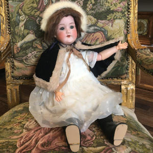 Antique doll with short brown hair in a white synthetic fibre dress and a black velvet cape and bonnet with fur trim. Sleep eyes.