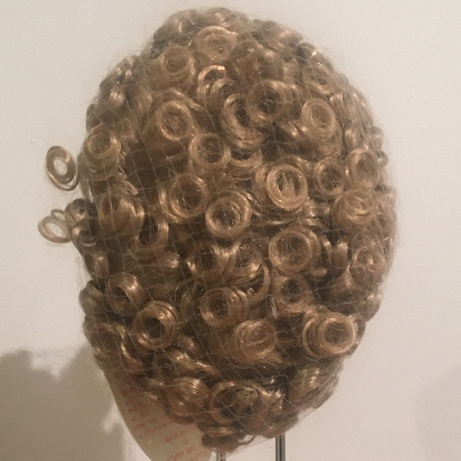 Light brown wig with short, tight curls, contained in a hair net, with a few curls protruding