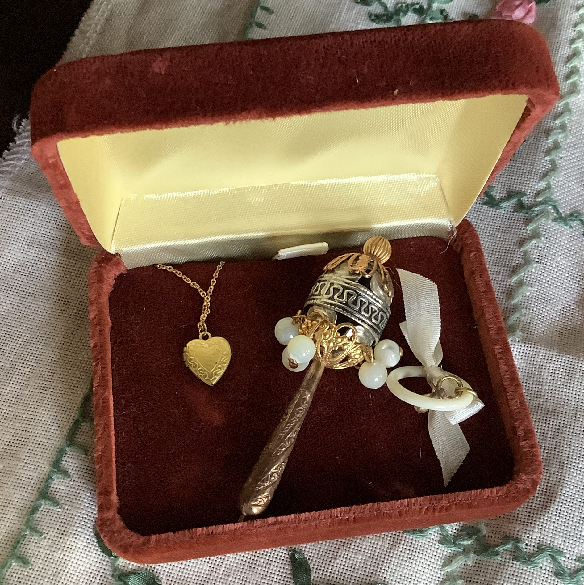 Burgundy velvet ring box containtin a gold- and silver-toned rattle, heart necklace, and pacifier