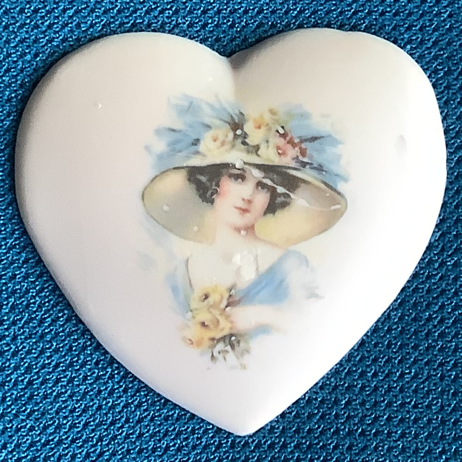 Heart-shaped porcelain ornament depicting a caucasian woman in a blue dress and large hat holding three yellow roses