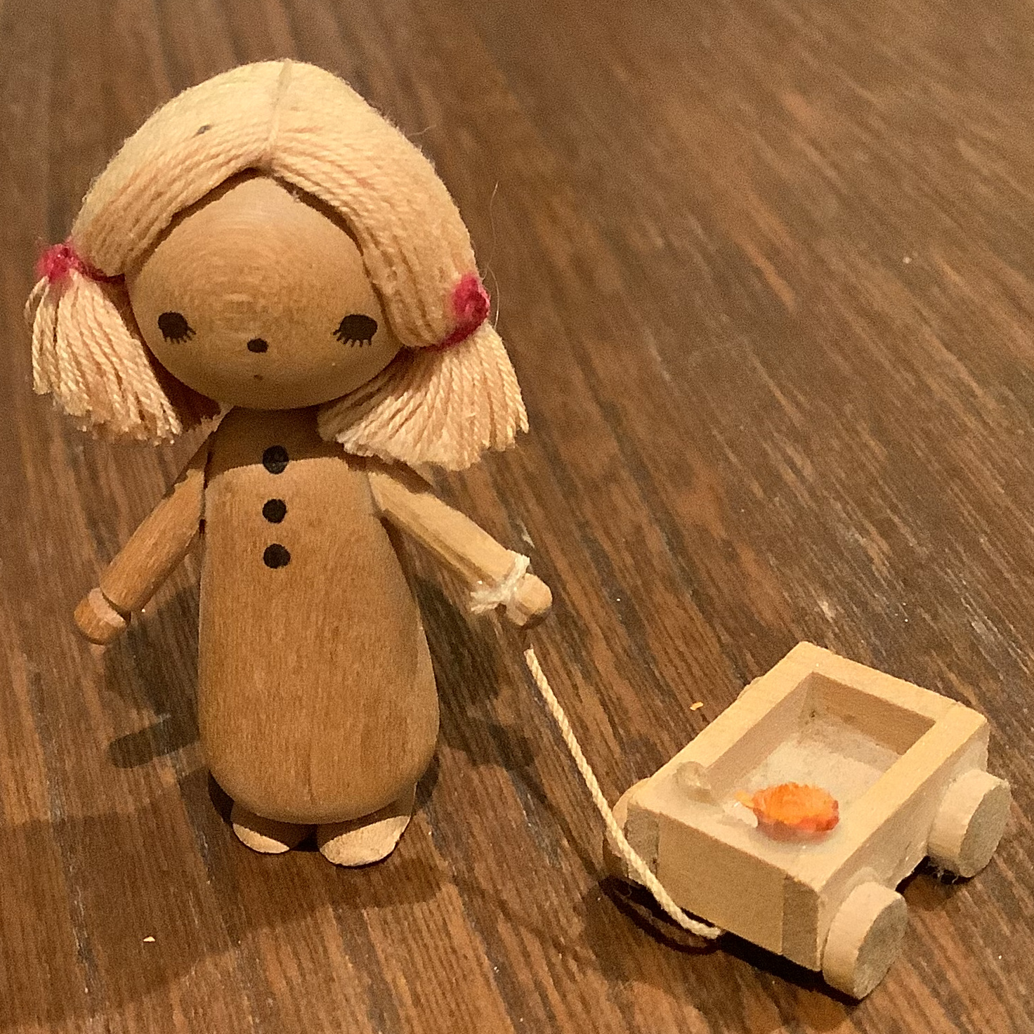 Minimalist wooden doll with yarn hair and painted face pulling a small wooden wagon containing a small, unidentifiable orange object