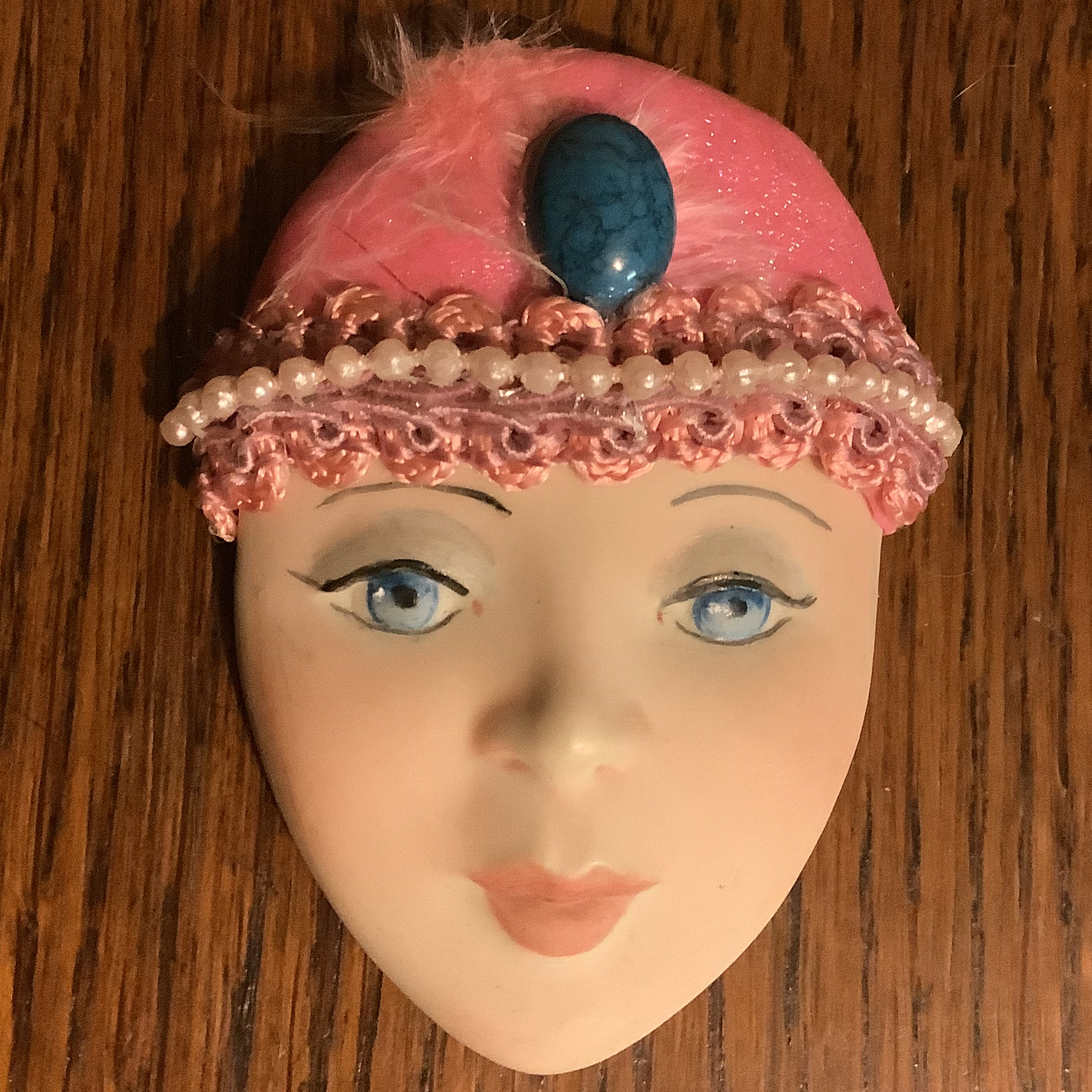 Brooch depicting a young, light-skinned, blue-eyed woman's face wearing a pink hat with beaded trim and large blue stone at forehead; visibly cracked