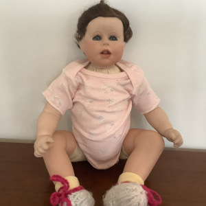Baby doll with porcelain head, arms and legs in frog posture with slightly annoyed facial expression, pink onesie