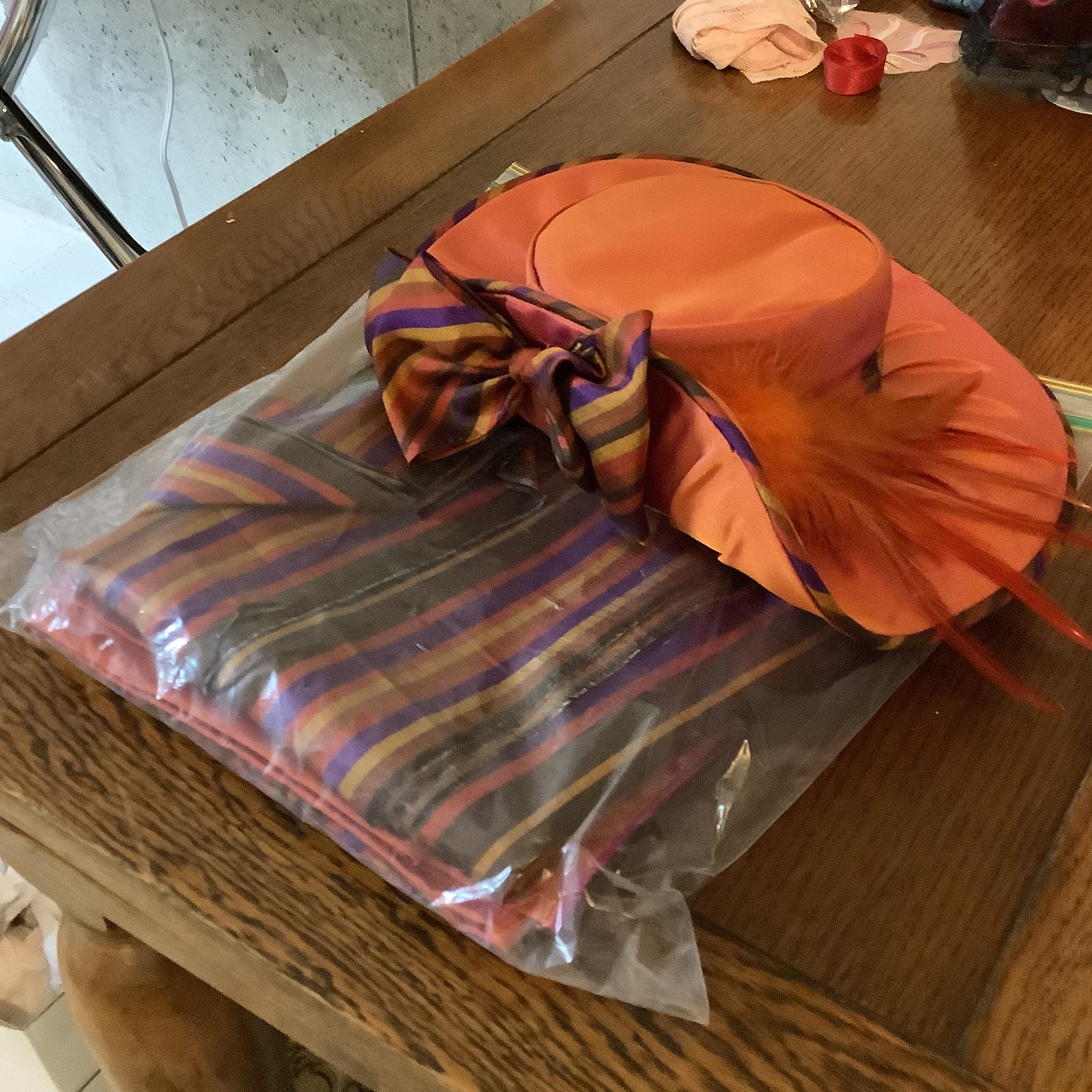 plastic bag containing striped orange and purple fabric, under a hat made from the same fabric with a bow and an orange feather