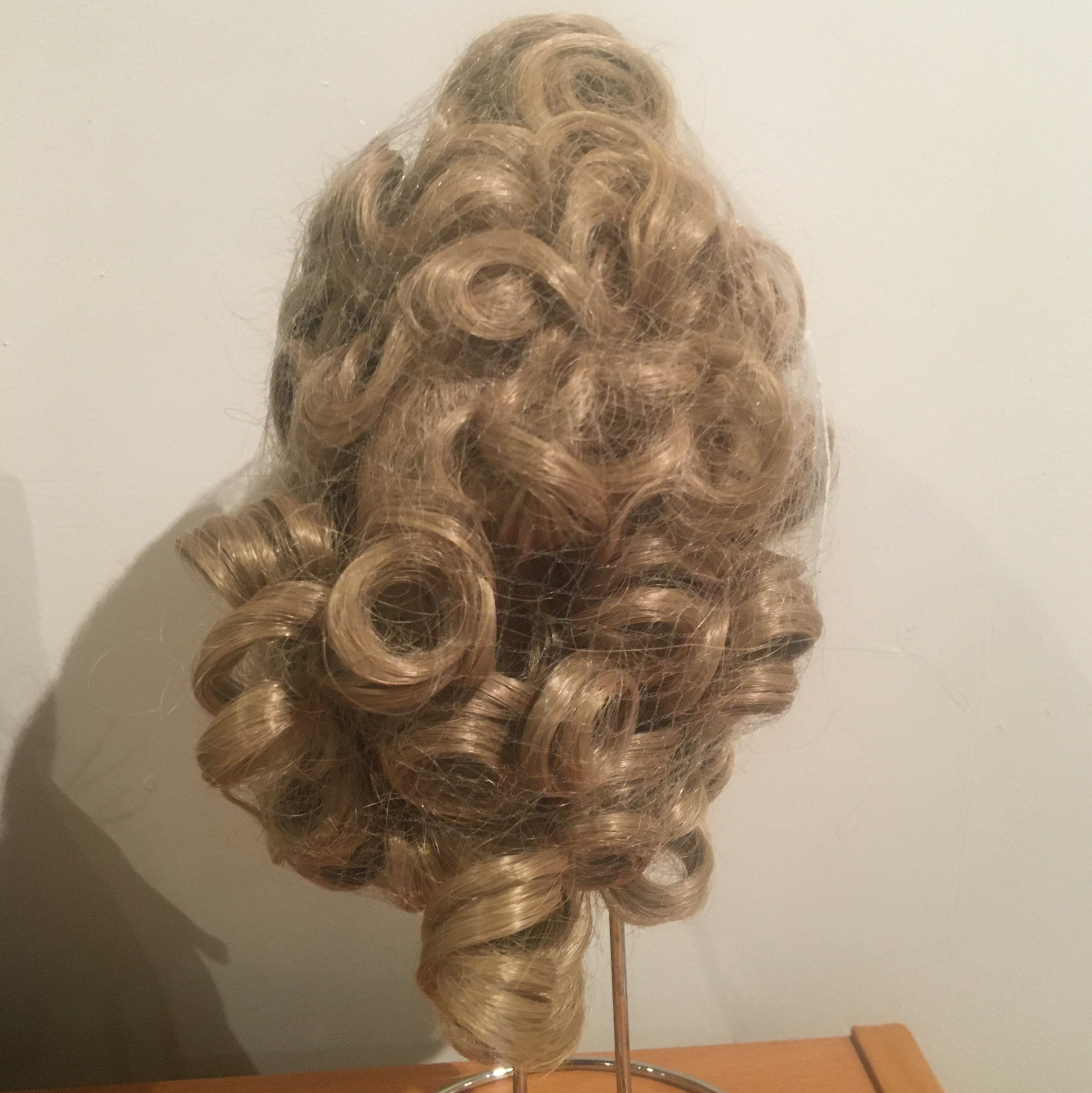 Back of blond wig, medium-length with large curls. Several curls protrude through a hole in a hair net