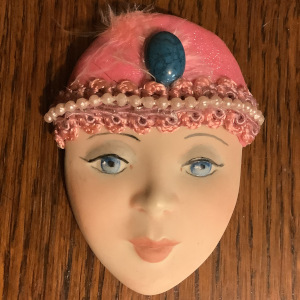 Brooch depicting a young, light-skinned, blue-eyed woman's face wearing a pink hat with beaded trim and large blue stone at forehead; visibly cracked