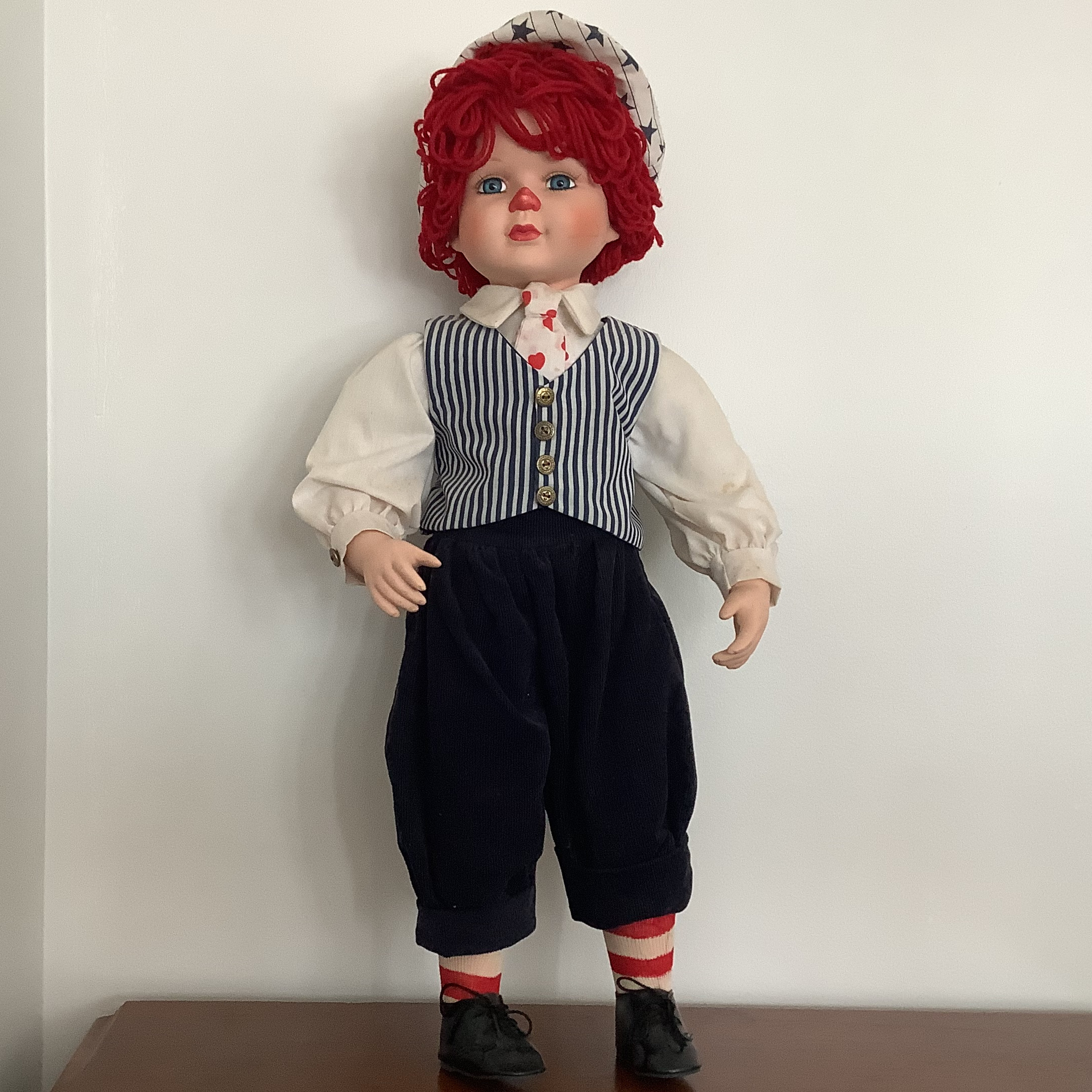 Reimagined Raggedy Andy doll with porcelain child's face painted to resemble the doll and curly yarn hair, with shit shirt, striped white and navy waistcoat, and navy trousers
