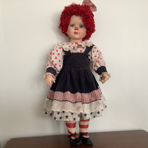 Reimagined Raggedy Ann doll with a child's face painted to resemble the doll, with short curly red hair and blue jumper dress with white patchwork dress underneath, printed in red and blue