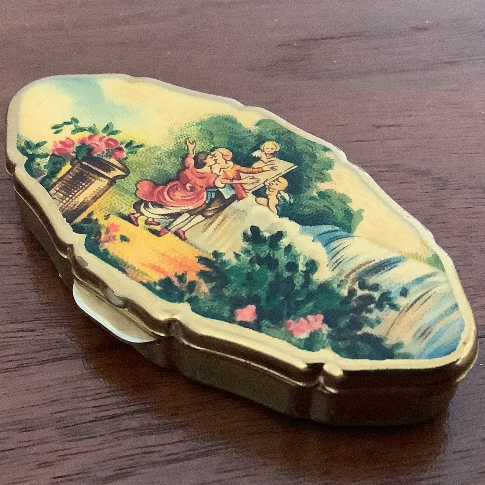 Closed pill box with gold-toned metal edges and pastoral scene printed on top