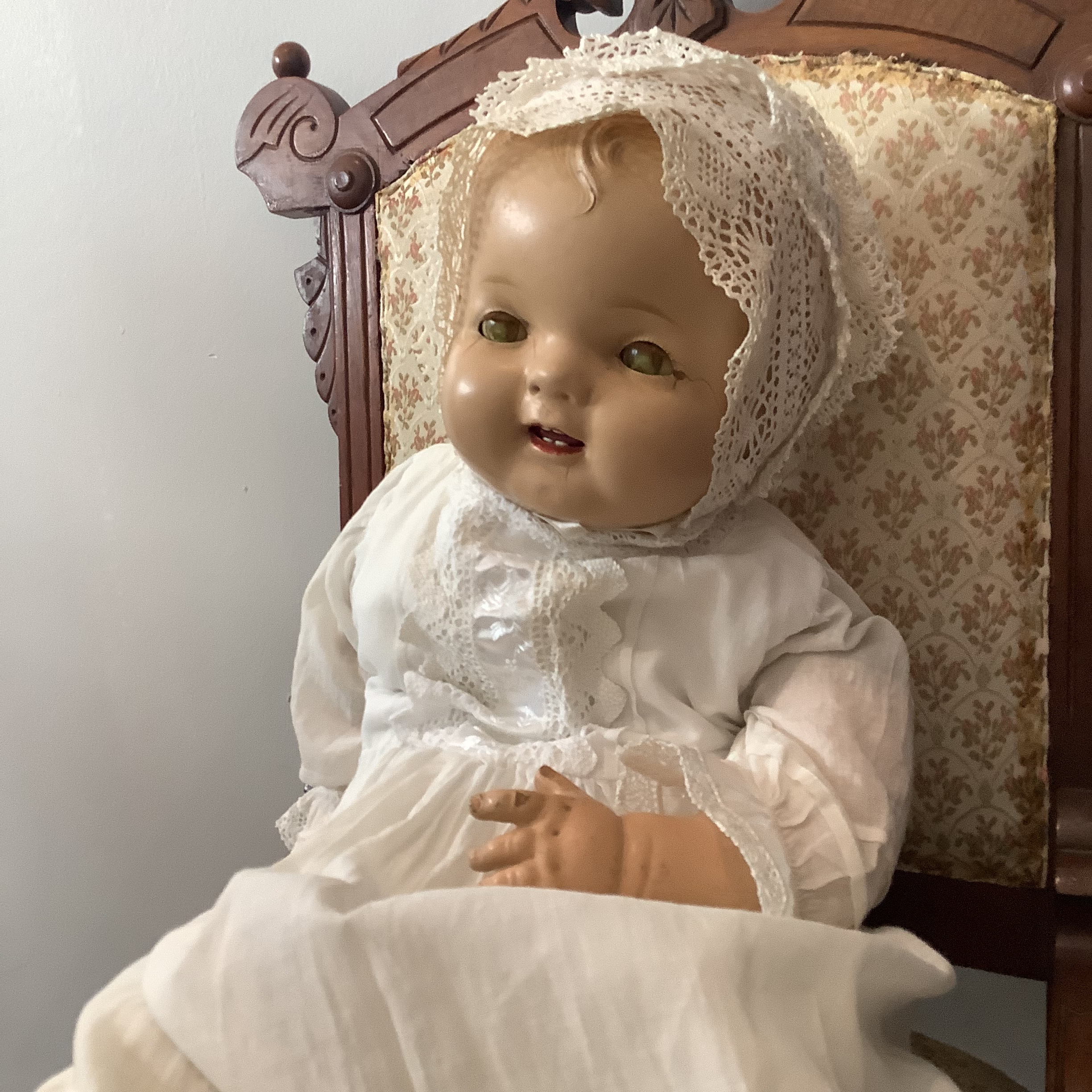 Vintage baby doll in elaborate christening gown with lace trim, seated in a chair.