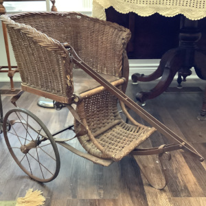 Brown wicker baby carriage with metal wheels, pull handle in front and broken leather seatbelt
