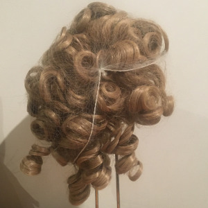 Back of blond wig, medium-length with large curls. Several curls protrude through a hole in a hair net