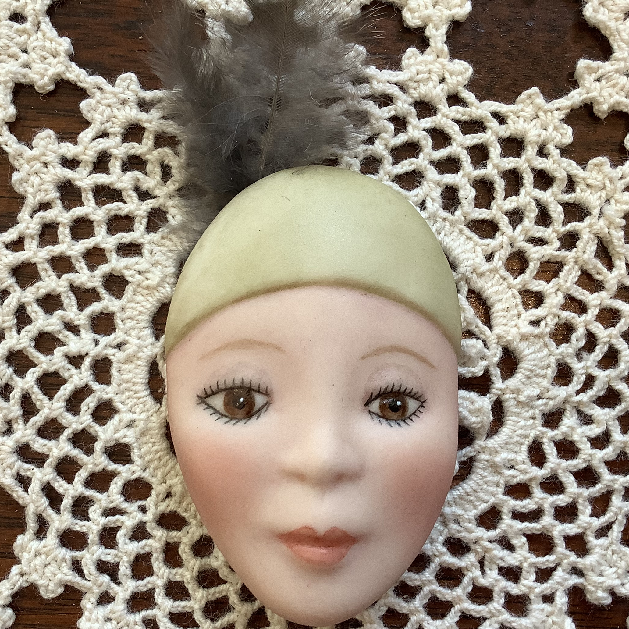 Brooch of a young, light-skinned woman's face, with heavy makeup and a green hat with a real feather
