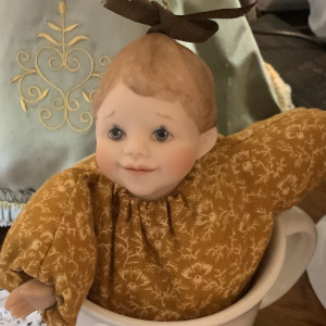 Small, light-skinned, orange-bodied half-doll with plush pillow body in coffee mug. Porcelain, handpainted hands and face with painted hair.